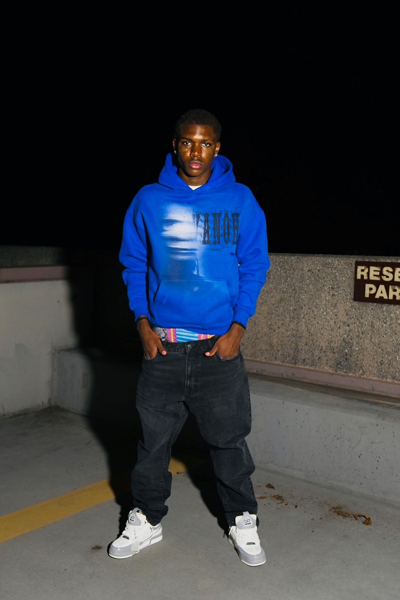 Model wearing a blue VANOH cropped hoodie with the ghost and VANOH print, black jeans and white sneakers, photographed at night
